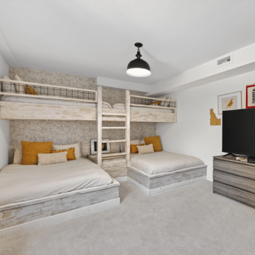 Modern bedroom with two sets of bunk beds, each with a lower double bed and upper single bed. The beds have neutral bedding and yellow accent pillows. A TV sits on a dresser across from the beds.