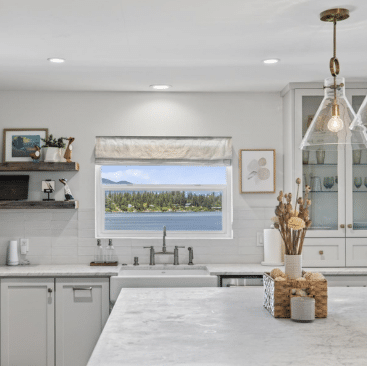 Bright, modern kitchen with white cabinets, marble countertops, open shelves, and decorative items. A large window above the sink offers a scenic view of a lake, trees, and mountains. A pendant light hangs above the island.