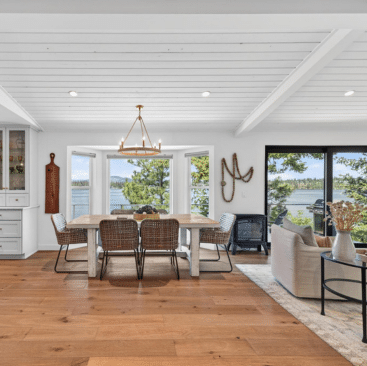 Open-concept dining and living area with large windows and sliding doors overlooking a lake, featuring a wooden dining table, wicker chairs, light wood flooring, and modern decor in neutral tones.
