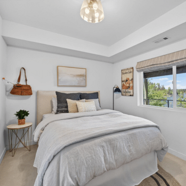 Bright bedroom with a large bed, neutral bedding, pillows, wall art, a small plant on a side table, and a window with a view of greenery. A pendant light hangs from the ceiling.