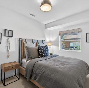 A cozy bedroom with a wooden bed frame, gray bedding, and pillows. A small wooden nightstand with a lamp is beside the bed. A window lets in natural light, and framed art decorates the white walls.