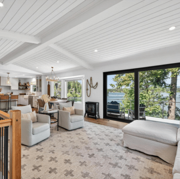 Bright, modern living room with white ceiling and walls, large windows overlooking a lake, cozy seating, a patterned rug, and a kitchen with bar stools in the background. Trees and water are visible outside.