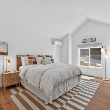 A bright, modern bedroom with a neatly made bed, striped rug, wooden nightstand with lamp, floor lamp, and large window letting in natural light. The decor features neutral tones and simple, cozy accents.