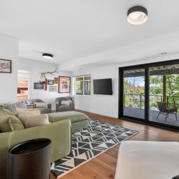 Modern living room with green and beige sofas, geometric rug, wall art, and large sliding glass doors opening to a balcony with a scenic outdoor view of trees and water. Wood floor and white walls give an airy feel.