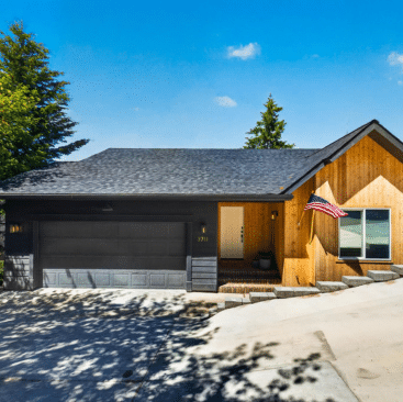 A modern single-story house with light wood siding, a dark gray garage, and a sloped driveway—ideal for vacation rental property management in North Idaho. An American flag is near the entrance, set among tall trees and a clear blue sky.