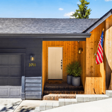 A modern house exterior with dark and natural wood siding, a white front door, potted plants by the entrance, an American flag, and the number 3711 displayed beside the garage—ideal for vacation rental property management in North Idaho.