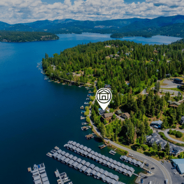 Aerial view of a forested lakeshore in North Idaho with houses, docks, and boats. A digital house icon marks a vacation rental property management client near the shoreline, surrounded by trees and mountains under a blue sky with clouds.