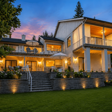Two-story modern house with large windows and balconies, warmly lit at dusk. Outdoor seating areas and a landscaped lawn are in front, with trees and a blue and pink sunset sky in the background—an inviting example of North Idaho vacation rentals perfect for those seeking comfort and style near Coeur d’Alene.